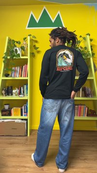 Μαύρο μακρυμάνικο πουκάμισο με γραφικό κύμα και αετό στην πλάτη, σε συνδυασμό με ανοιχτό μπλε φαρδιά τζιν, μπροστά σε κίτρινο τοίχο.