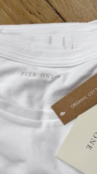 T-shirt branca de algodão orgânico com uma textura suave, apresentando a impressão "PIER ONE" nas costas. Acompanha uma etiqueta castanha e creme.