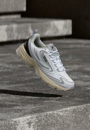 Hvid og beige Fila-sneakers med sorte detaljer og snørebånd, svæver over en tekstureret betonoverflade med trapper i baggrunden.