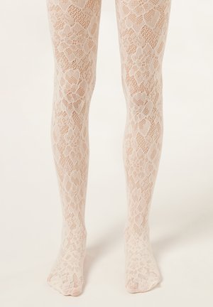 Ben iført beige strømpebukser med blondermønster med blomstret og geometrisk design, stående på en lys baggrund.