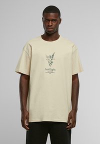 Beige oversized t-skjorte laget av bomull, med et grønt grafisk trykk av en engel og teksten "Lucid Lights" i midten.