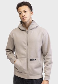 Haut à capuche zippé beige clair avec capuche, deux poches avant, poignets côtelés et une étiquette tissée sur la poche. Tissu doux en mélange de coton.