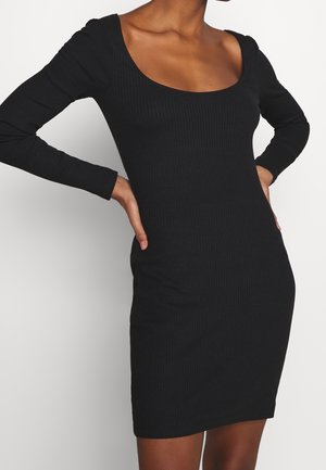 Robe noire moulante à côtes avec des manches longues, décolleté arrondi et une silhouette ajustée qui met en valeur la forme.