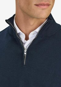 Laivastonsininen vetskarihuppari, jossa on korkea kaulus, valmistettu neulotusta materiaalista; siinä on hopeanvärinen vetskari ja se on kerroksittain valkoisen kauluspaitan päällä.