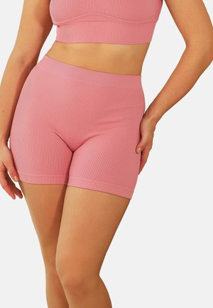 Frau trägt gerippte, pinke Biker-Shorts mit hohem Bund und passendes Crop-Top, eine Hand auf der Hüfte ruhend.