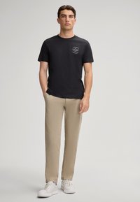 T-shirt en coton noir avec un petit graphique blanc sur la poitrine gauche, associé à un pantalon beige et des baskets blanches. Design simple et décontracté.