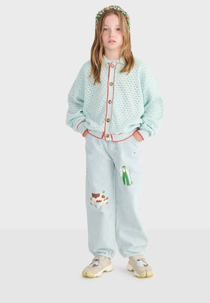 Dziewczyna z długimi włosami nosi jasnoniebieski dziergany kardigan, jasne dżinsy z nadrukiem farmera i krów, kwiatową opaskę na głowę oraz trampki z motywami zwierzęcymi.