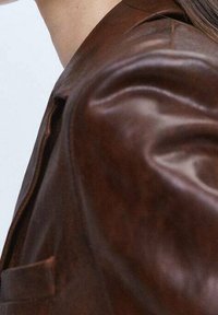 Ruskea nahkatakki, jossa on sileä pinta, jämäkkä olkapään muotoilu ja lovikaulus. Siinä on yksi rintatasku ja minimalistinen muotoilu.