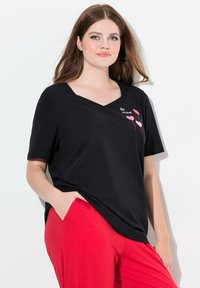 Zwarte v-hals t-shirt met roze ballonafbeeldingen en de tekst "Be mine." Gecombineerd met rode broeken, wat een casual silhouet laat zien.