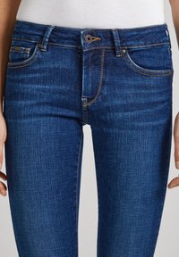Jeans de denim azul oscuro con un diseño ajustado, que presentan cinco bolsillos, costuras doradas y un cierre de botón metálico. Textura suave.
