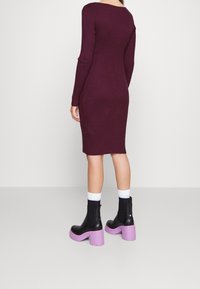 Persona con un vestido ajustado de manga larga y con canalé en color púrpura, que lleva botas negras hasta el tobillo con gruesos tacones púrpuras y calcetines blancos.