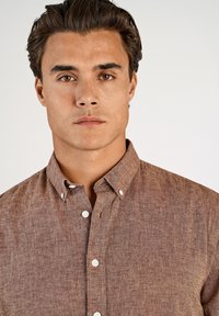 Bruine overhemd met knopen gemaakt van gestructureerde stof, voorzien van een kraag, witte knopen en een borstzak.