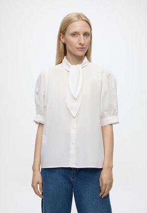 Femme aux cheveux blonds et raides, portant un chemisier blanc à manches courtes bouffantes et un nœud à l'avant, associée à un jean bleu, debout devant un fond uni.