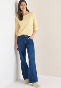 Pull léger à rayures jaunes avec manches 3/4, associé à un jean en denim bleu à jambes larges. Chaussures : baskets légères. Fond : mur uni.