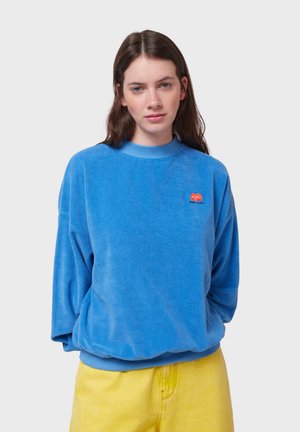 Sweatshirt en velours bleu clair avec un col rond côtelé, des épaules tombantes et un logo en forme de cœur rouge sur la poitrine. Associé à un short jaune.