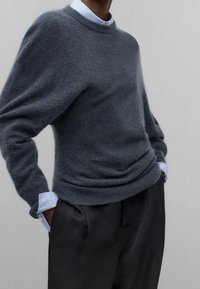 Donkergrijze wollen trui met een ronde hals, losse pasvorm en geribbelde boorden, gelaagd over een lichtblauw gestreept overhemd en zwarte broek.