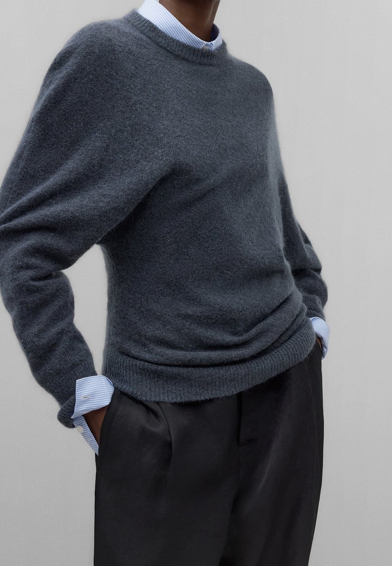 Donkergrijze wollen trui met een ronde hals, losse pasvorm en geribbelde boorden, gelaagd over een lichtblauw gestreept overhemd en zwarte broek.