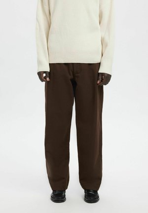 Person iført en cremefarvet ribstrikket sweater, løstsiddende mørkebrune bukser og sorte loafers, stående mod en ensartet lys baggrund.