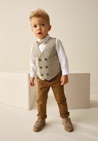 Enfant portant un gilet à carreaux clair avec des boutons marron, une chemise blanche et un nœud papillon beige. Pantalon marron et chaussures grises à lacets avec des accents.