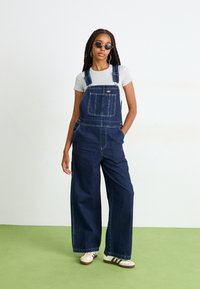 Salopette in denim blu scuro con gambe larghe, tasche frontali e bretelle regolabili, indossata su una maglietta grigia. Accompagnata da occhiali da sole e sneaker.