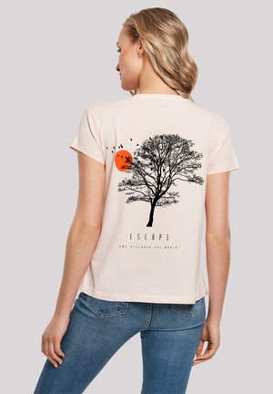 T-shirt rose clair avec un graphique d'arbre noir et un soleil orange, surplombé d'oiseaux. Le texte en dessous dit "ÉCHAPPEZ-VOUS ET DÉCOUVREZ LE MONDE."