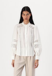 Weiße Bluse mit aufwendiger Spitzenborte und aufgeblasenem Ärmel. Sie hat einen hohen Ausschnitt und eine Knopfleiste vorne, kombiniert mit beigen Hosen.