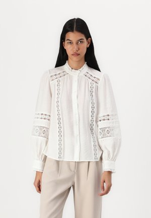 Hvit bluse med intrikate blonder og puffede ermer. Har høy hals og knapper foran, kombinert med beigefargede bukser.