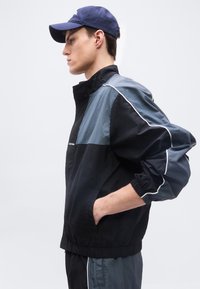 Νέος άνδρας με μαύρο και γκρι αθλητικό μπουφάν και αντίστοιχο παντελόνι, φοράει μπλε καπέλο μπέιζμπολ, ποζάροντας προφίλ με τα χέρια στις τσέπες του μπουφάν.