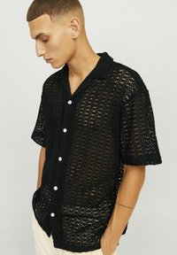 Czarna, ażurowa koszula z lekkiej, oddychającej tkaniny. Krótkie rękawy i zapięcie na guziki z białymi guzikami. Teksturowany wzór na całej powierzchni.