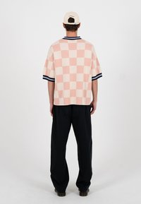 Bărbat purtând o cămașă cu mâneci scurte, în carouri roz și crem, pantaloni negri și o șapcă bej, stând cu fața spre departe pe un fundal simplu.