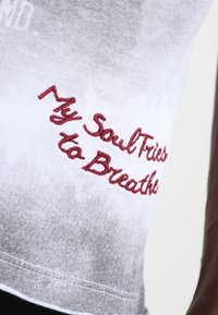 Hvid og grå gradient tanktop med rød broderet tekst, der står "Min Sjæl Forsøger at Ånde." Stoffet har en blød tekstur.