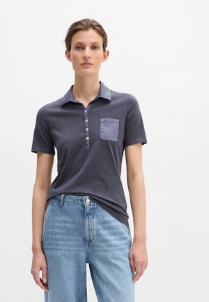 Frau trägt ein dunkles kurzärmeliges Poloshirt mit Knöpfen und einer Brusttasche, kombiniert mit hellblauen, hoch taillierten Jeans.