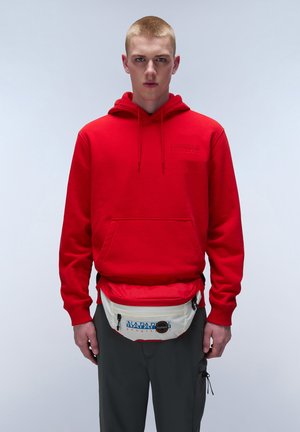 Red hoodie with a front pocket and drawstrings. White fanny pack with blue logo and black accents. Gray pants. Simple background.