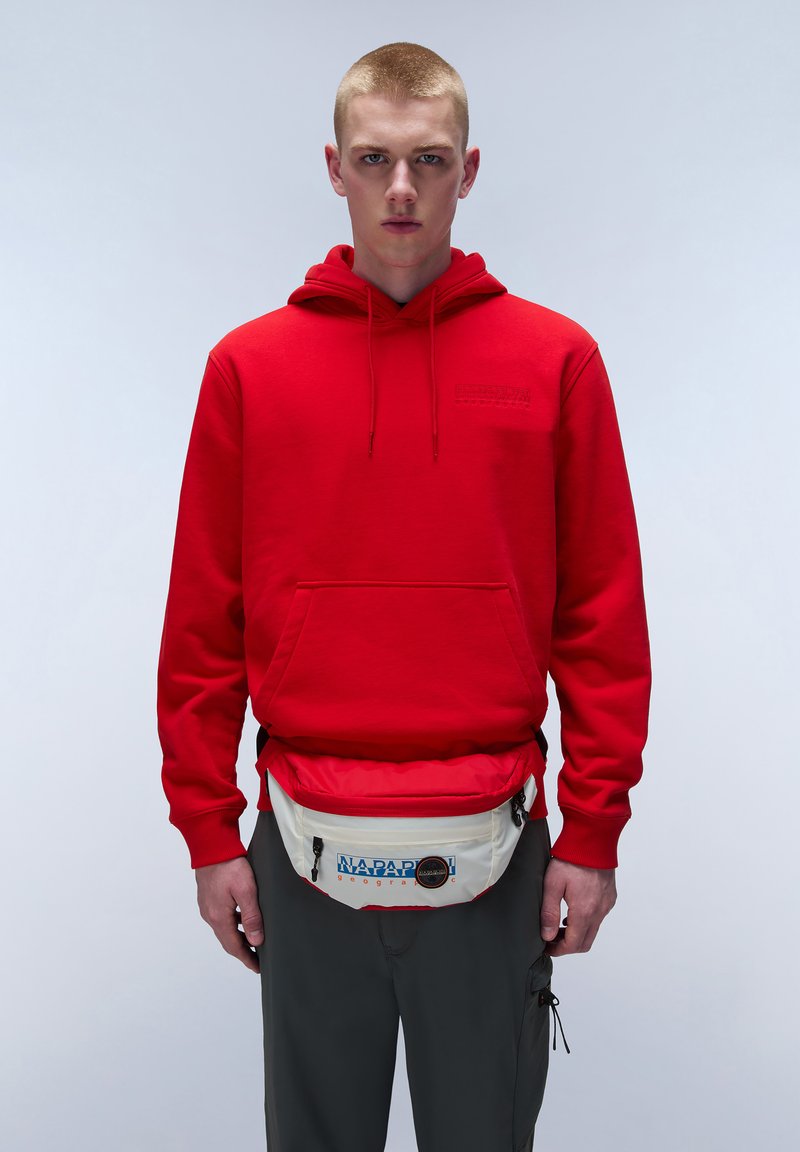 Sudadera roja con bolsillo frontal y cordones. Riñonera blanca con logo azul y detalles negros. Pantalones grises. Fondo simple.