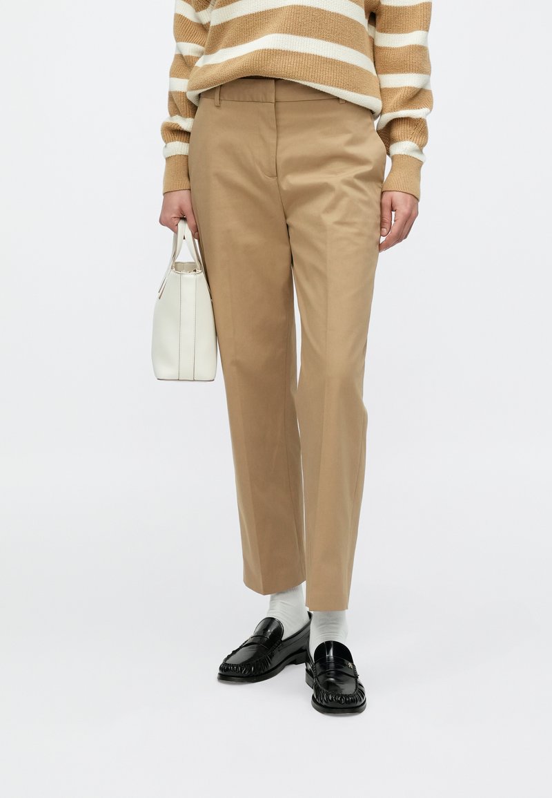 Bež chinos hlače s prilagođenim krojem, glatke teksture, uparene s crnim loafericama i malom bijelom torbicom.
