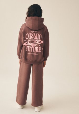 Osoba ubrana w brązową bluzę z kapturem i spodnie oraz białe trampki, napis na bluzie to "Vision of a Cosmic Future" z motywem planety i gwiazd.