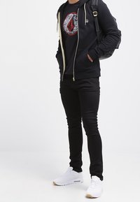 Chaqueta con cremallera negra y detalles en crema, camiseta gráfica negra, pantalones negros ajustados y zapatillas blancas con suelas de goma. Mochila al hombro.