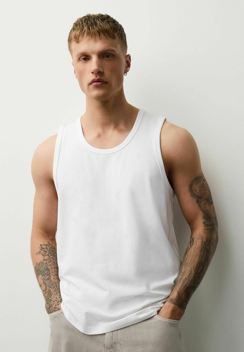 Jeune homme aux cheveux blonds courts portant un débardeur blanc, un pantalon beige et des tatouages visibles sur les deux bras, sur un fond uni.