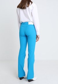 Un pantalon évasé bleu en tissu lisse et extensible. Le design comprend une ceinture avec une étiquette et deux poches arrière. Porté avec une chemise blanche.