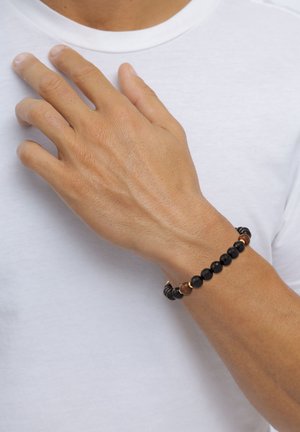 Perlenarmband mit schwarzen und braunen Perlen in glatter Optik, am Handgelenk vor einem weißen Hemd-Hintergrund.
