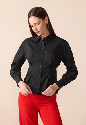 Chemise noire à manches longues avec un col, fermeture à boutons sur le devant et des détails plissés ; le tissu semble lisse et légèrement brillant.