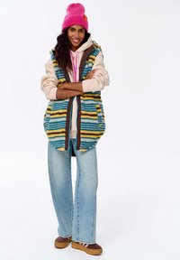 Erksinine, triibuline vest, millel on sinised, kollased ja pruunid toonid, koos heleda kapuutsiga dressipluusiga ja laia jalaga heleda pesu teksadega.