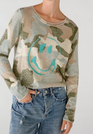 Person iført en camouflage-langærmet sweater med et stort turkis smilende ansigt-design og blå slidte jeans med en hånd i lommen.