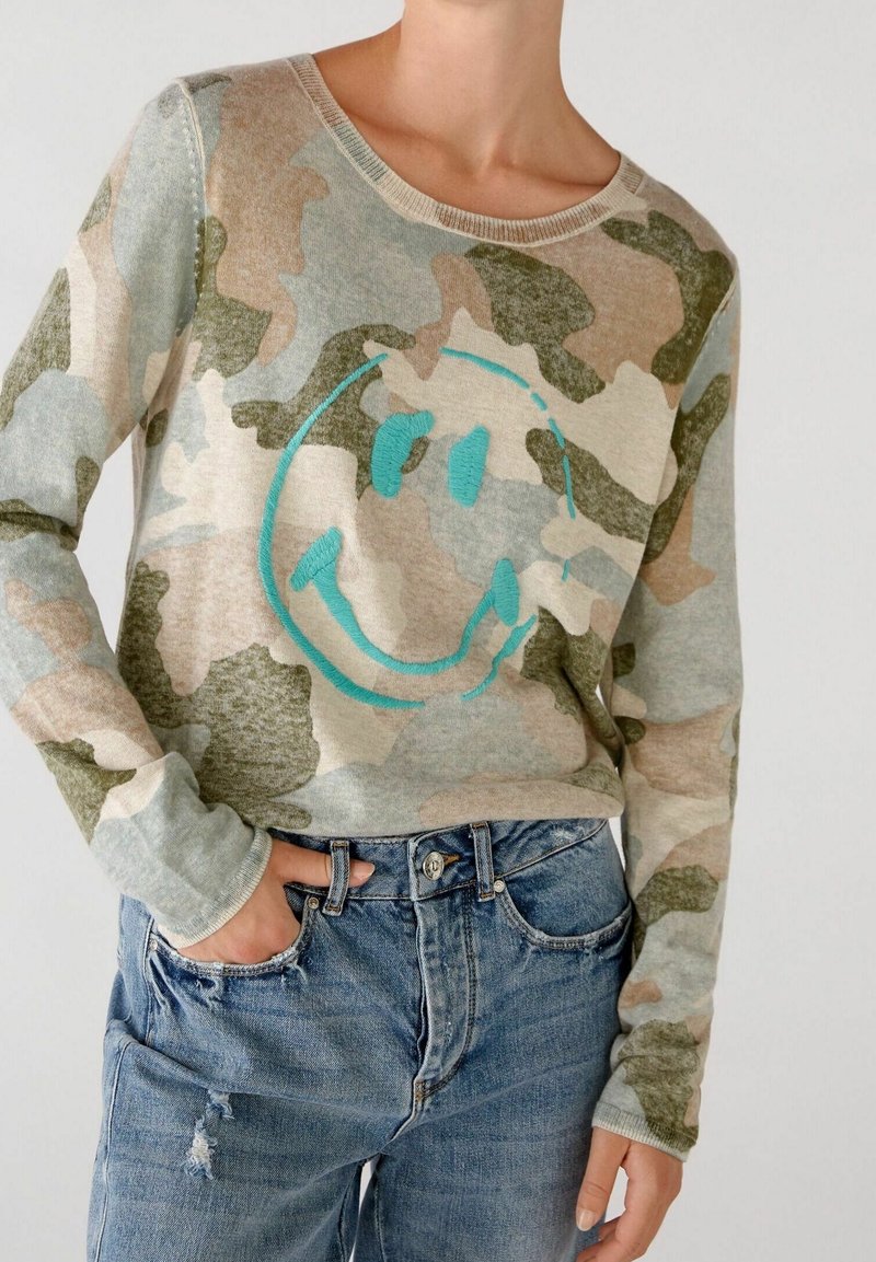 Personne portant un pull à manches longues camouflage avec un grand motif visage souriant turquoise et un jean bleu déchiré avec une main dans la poche.