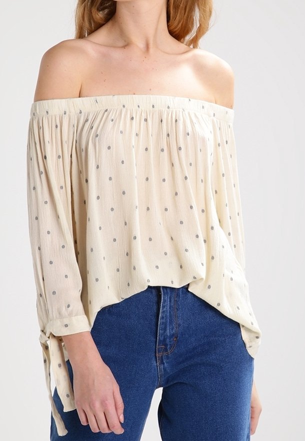 Femme portant un chemisier crème épaules dénudées avec de petits pois et des liens aux manches, associé à un jean bleu taille haute.
