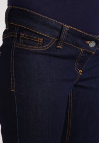 Tmavě modré džínové kalhoty s kontrastním oranžovým prošitím, standardními kapsami a kovovým zapínáním. Hladká textura a přiléhavý střih.