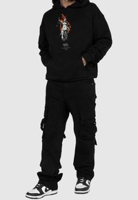 Felpa nera con cappuccio e stampa grafica di una statua che tiene una bilancia e fiamme. Abbinata a pantaloni cargo neri con tasche laterali e cinturini. Scarpe da ginnastica bianche.