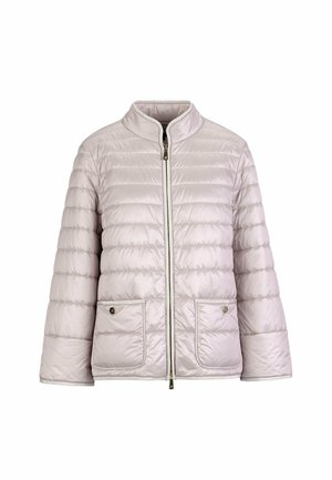 Woman wearing a light pink quilted jacket over a scarf, white pants, beige heels, and a gold bracelet, standing against a white background.