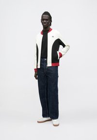 Chaqueta bomber blanca y negra con detalles en rojo, usada sobre una camiseta negra. Combinada con jeans de mezclilla oscuros y sueltos y zapatillas blancas con suelas marrones.