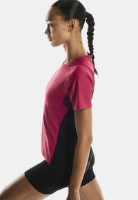 Sportovní outfit s jednobarevným růžovým tričkem s krátkým rukávem a černými postranními panely, doplněný o černé šortky. Materiál se zdá být lehký a prodyšný.